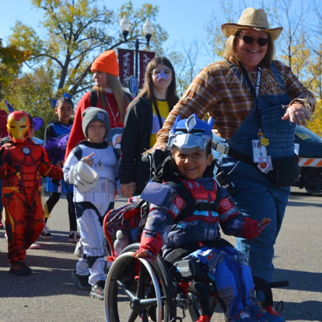 Student in costume and wheelchair smiles with teacher walking down street in parade